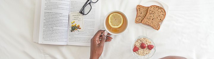 A birds-eye view of a black person's crossed legs as they sit in bed with breakfast and a book